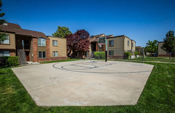 A basketball court is located in the middle of a grassy area in front of apartment buildings.